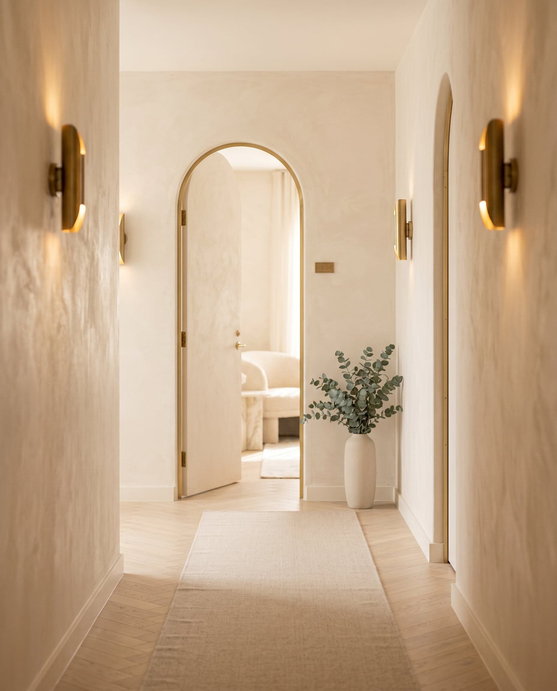 Private suite hallway with cream herringbone floor, brass sconces, and an arched doorway leading to a suite.