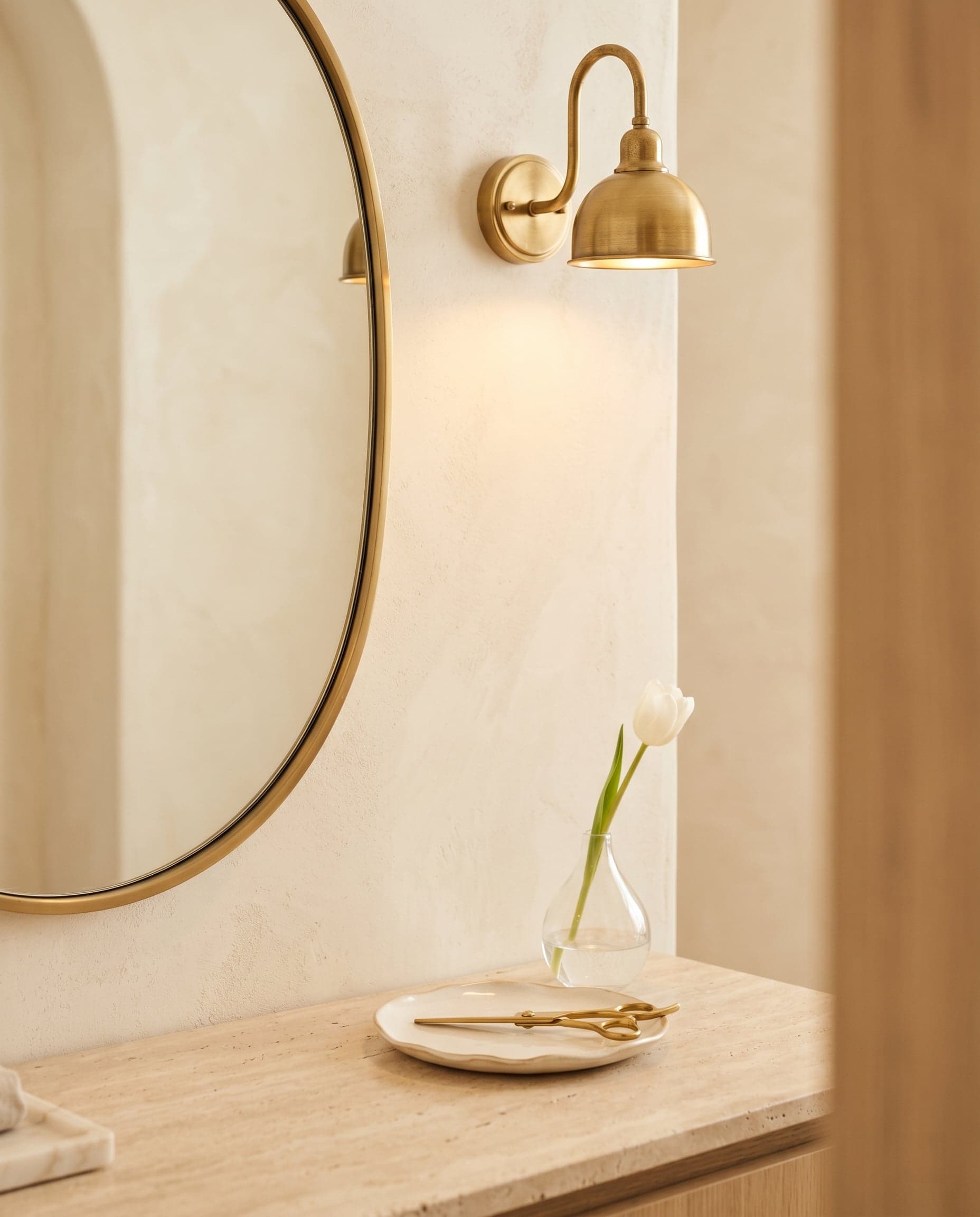Close-up of a styling station: brass gooseneck lamp, oval mirror, ceramic dish with brass-handled scissors, single white tulip.