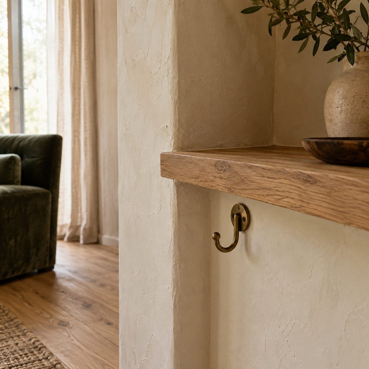 Corner of cream lime-plaster wall meeting an oak shelf, with a single brass hook and a green velvet chair beyond.